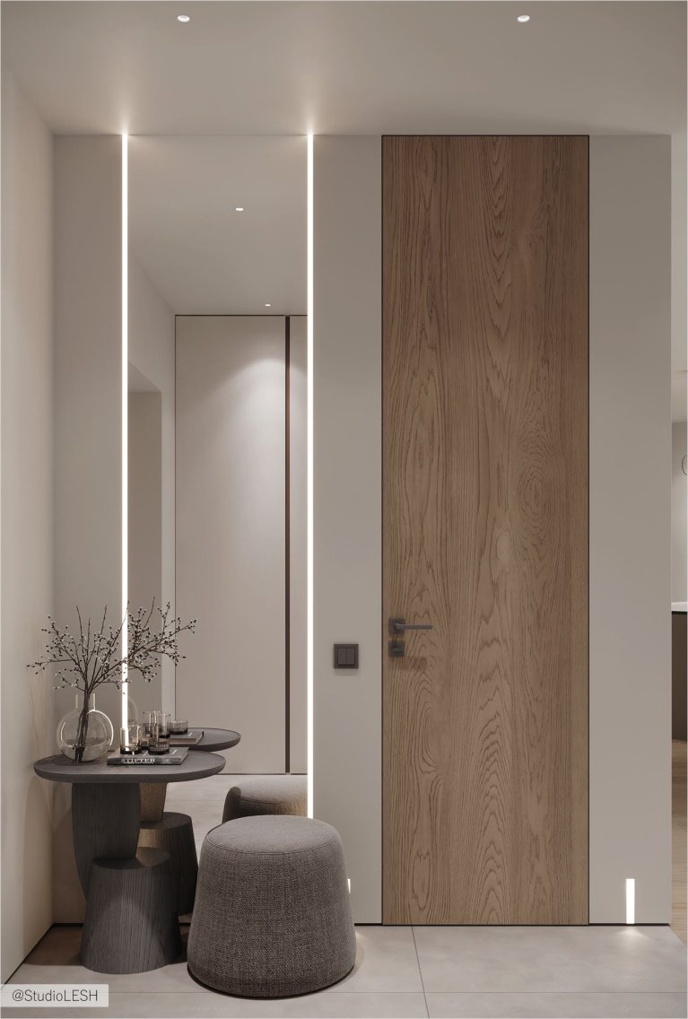 Современная прихожая в минималистичном стиле с деревянной входной дверью, высоким зеркалом с LED-подсветкой и стильной мебелью. Компактный столик с декоративными элементами и мягкие пуфы создают уютную атмосферу. Светлые стены и встроенное освещение подчеркивают лаконичный дизайн