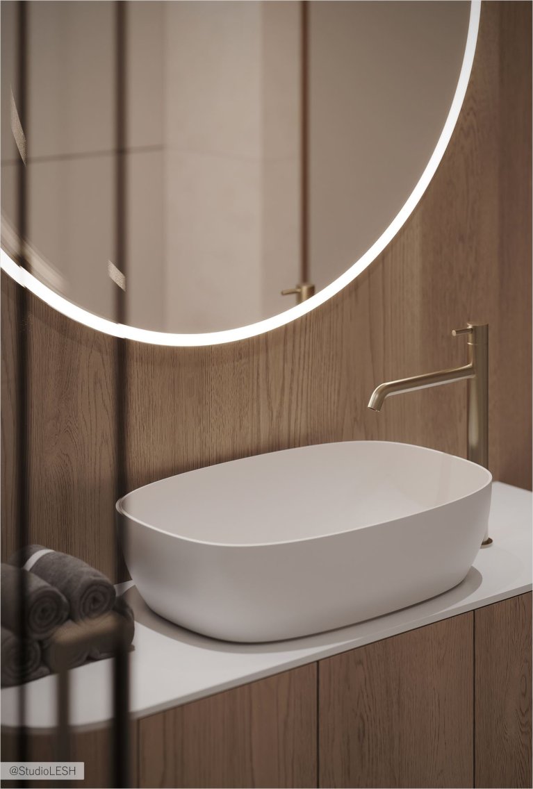 Современная ванная комната с овальной раковиной, установленной на белой столешнице. Зеркало с круговой подсветкой отражает минималистичный дизайн. На заднем плане видны деревянные панели, добавляющие текстуру и теплоту