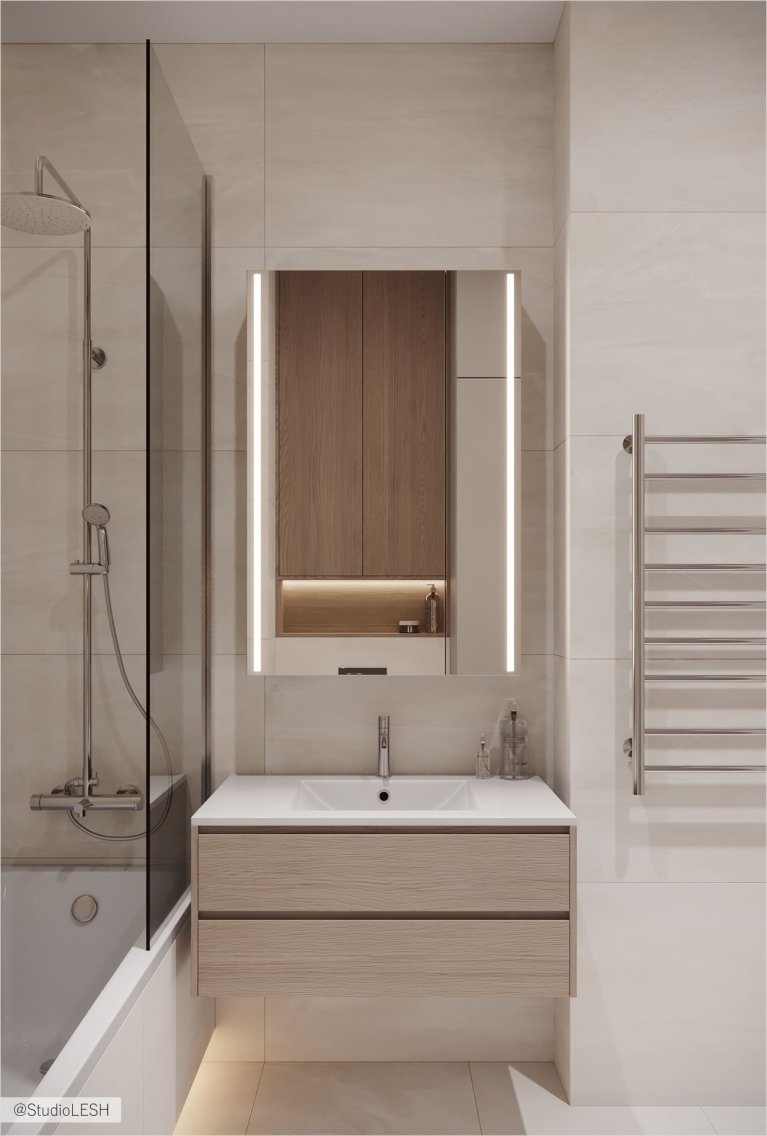 Интерьер ванной комнаты в светлых тонах с душевой кабиной, частично огороженной стеклом, зеркалом с подсветкой, деревянным шкафчиком и тумбой под раковину с двумя выдвижными ящиками. На заднем плане - полотенцесушитель