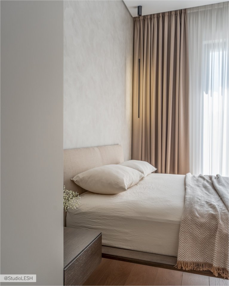 Уютная спальня с большой кроватью, застеленной серым покрывалом, и декоративными подушками. На стене за кроватью - деревянная панель, а по бокам - две тумбочки с лампами. В комнате также есть большое окно с жалюзи и плотными шторами, пропускающими мягкий свет