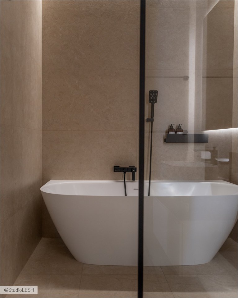 Стильная ванная комната с отдельно стоящей ванной с черным смесителем и душевой кабиной с тропическим душем и черной фурнитурой. На заднем плане - полочки с банными принадлежностями и зеркало с подсветкой