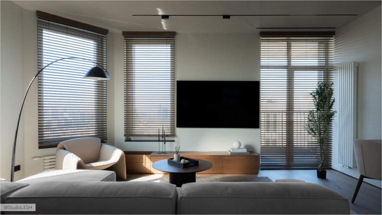 Уютная гостиная с мягким угловым диваном, украшенным подушками, и журнальным столиком с декоративными элементами. На заднем плане - высокие окна с деревянными жалюзи, пропускающими яркий солнечный свет, и комнатное растение