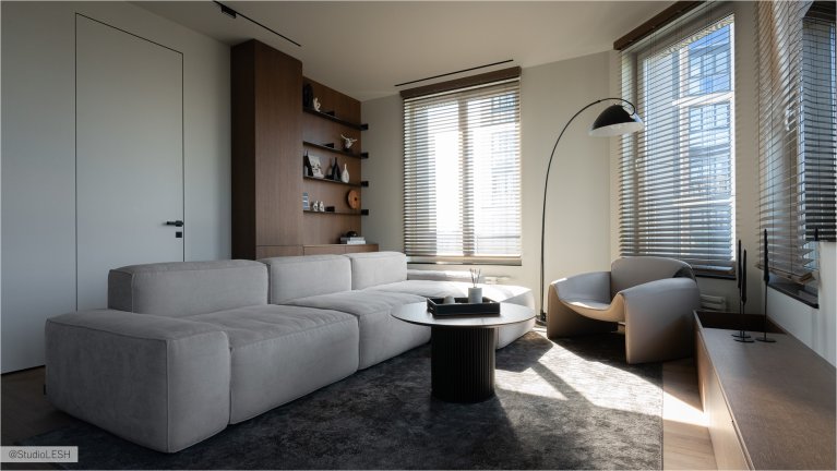 Интерьер гостиной в светлых тонах с серым модульным диваном, удобным креслом с мягкой обивкой, высокими окнами с деревянными жалюзи, плоским телевизором на стене и разнообразным декором, включая растения и светильники