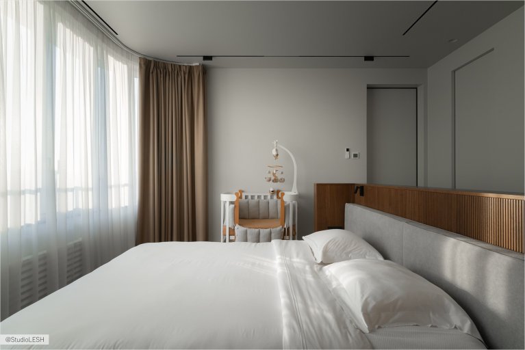 Интерьер спальни в светлых тонах с большой двуспальной кроватью, деревянной стеной за изголовьем, двумя тумбочками по бокам и подвесными светильниками. В комнате также есть большое окно с жалюзи и плотными шторами