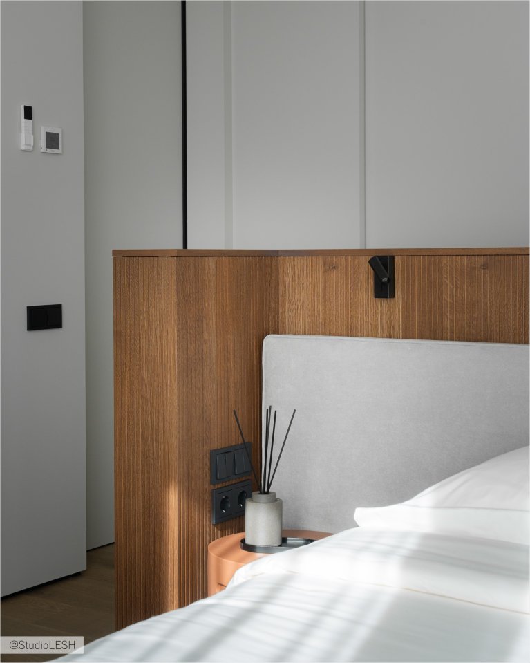 Интерьер спальни в светлых тонах с большой двуспальной кроватью, деревянной стеной за изголовьем, двумя тумбочками по бокам и подвесными светильниками. В комнате также есть большое окно с жалюзи и плотными шторами