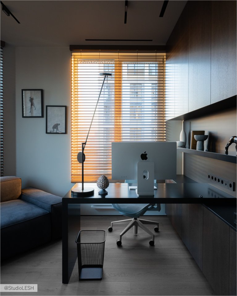 Стильный рабочий кабинет с большим письменным столом, на котором расположен компьютер Apple с большим монитором. Рядом с компьютером стоит настольная лампа и лежат различные предметы. На заднем плане - удобное эргономическое кресло и окно с жалюзи, пропускающими яркий солнечный свет