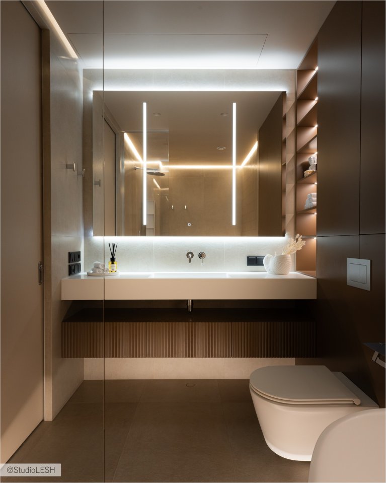 Интерьер ванной комнаты в светлых тонах с большой прямоугольной раковиной, установленной на деревянной тумбе с рифлеными фасадами. Над раковиной висит большое зеркало с подсветкой, отражающее светлые стены и полки с аксессуарами. Видны также черные смесители и электрические розетки