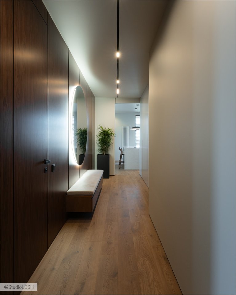 Интерьер коридора в светлых тонах с большим прямоугольным зеркалом с подсветкой, мягкой скамейкой с тканевой обивкой, встроенными шкафами с гладкими фасадами и паркетным полом. Слева виден небольшой столик с вазой и вешалкой