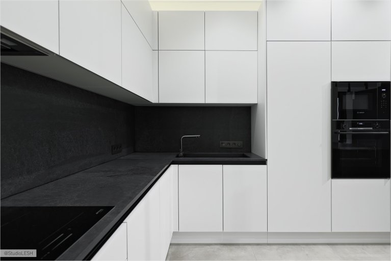 Стильная кухня с белыми шкафами с гладкими фасадами, черной столешницей, на которой расположены мойка и варочная поверхность. Встроенная техника гармонично сочетается с общим дизайном кухни. Освещение играет важную роль в создании уютной атмосферы