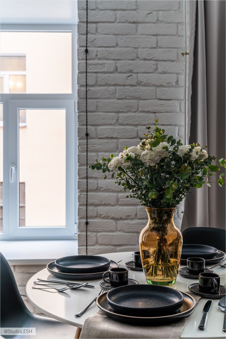 Элегантный обеденный стол с темной посудой, украшенный букетом белых цветов в янтарной вазе, создает уютную атмосферу в светлой столовой